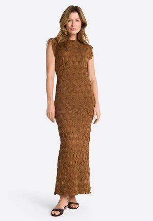 Donna con capelli mossi che indossa un abito lungo marrone senza maniche e con texture, e sandali neri aperti davanti, in piedi davanti a uno sfondo chiaro e uniforme.