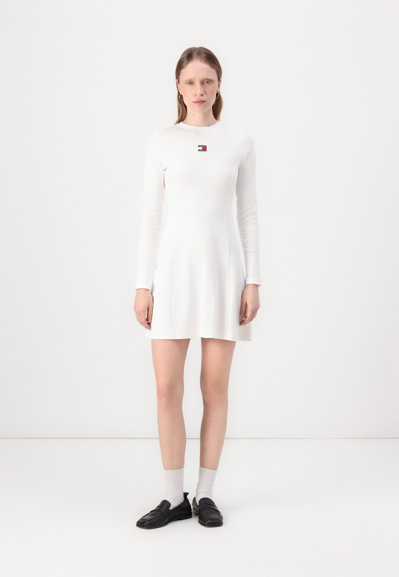White long-sleeve dress with a fitted bodice and flared skirt. Features a small logo in red and navy. Worn with black loafers and grey socks.