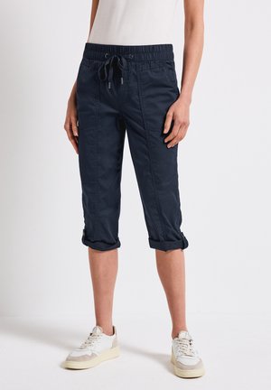 Person iført mørkeblå cropped bukser med snøre og opkrogede manchetter samt hvide sneakers, stående mod en ensfarvet baggrund.