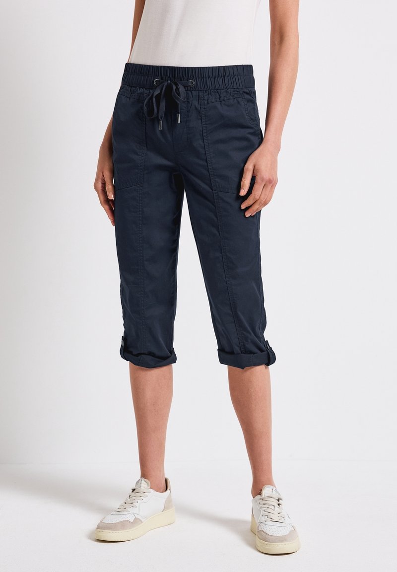 Personne portant un pantalon court bleu foncé avec cordon et revers roulés, ainsi que des baskets blanches, debout devant un fond uni.