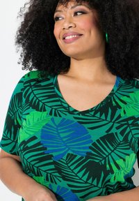 Grön kortärmad t-shirt med V-hals, med ett tropiskt lövtryck i nyanser av grönt och blått på en mörk bakgrund.