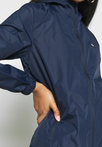 Granatowa kurtka przeciwwiatrowa wykonana z lekkiego, błyszczącego materiału, z zamkiem błyskawicznym z przodu, elastycznymi mankietami i bocznymi kieszeniami.