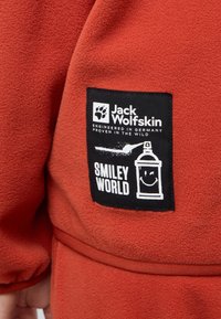 Garment in pile rosso con un'etichetta nera con testo e grafica bianchi, che recita "Jack Wolfskin" e "SMILEY WORLD," raffigurante una bomboletta spray sorridente.