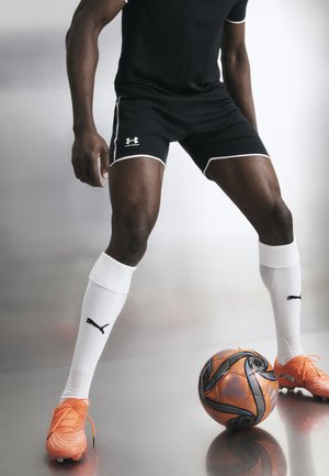 Αθλητής με μαύρα σορτς, λευκές κάλτσες Puma και πορτοκαλί ποδοσφαιρικά παπούτσια στέκεται έτοιμος με μια πορτοκαλί και μαύρη ποδοσφαιρική μπάλα σε αντανακλαστικό δάπεδο.