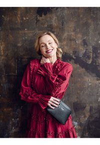 Červené volánkové šaty s dlouhými rukávy a texturovanou látkou, doplněné malou černou kabelkou přes rameno s elegantním kováním a vyraženým logem.