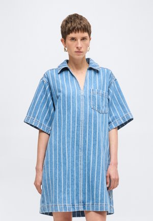 Žena s krátkymi vlasmi oblečená v modro-biele denimové šaty s vertikálnymi pruhmi, krátkymi rukávmi a náprsným vreckom, stojaca pred jednofarebným pozadím.