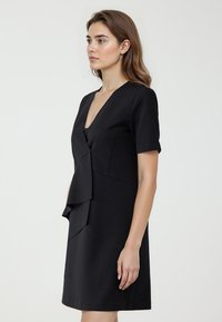 Robe noire avec un décolleté en V et des manches courtes. Présente un design avant superposé et une texture lisse. Coupe simple, longueur au genou.