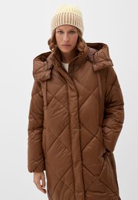 Bruine gewatteerde pufferjas met een capuchon, met diagonale stiksels en trekkoorden. Gecombineerd met een gele en bruine gestreepte gebreide muts.