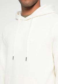 Biała bluza z kapturem z kieszenią typu kangur, sznurkami i subtelnym trójkątnym logo. Miękka tkanina o gładkiej fakturze i luźnym kroju.