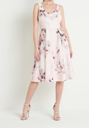 Kobieta ubrana w bezrękawową, sięgającą kolan, bladoróżową sukienkę w kwiaty z dopasowanym gorsetem i rozkloszowaną spódnicą, połączoną ze srebrnymi sandałami na wysokim obcasie.