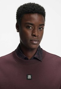 Maroon sweatshirt with round neckline, textured fabric, and a black HU GO logo patch. Layered with a dark patterned collared shirt underneath.