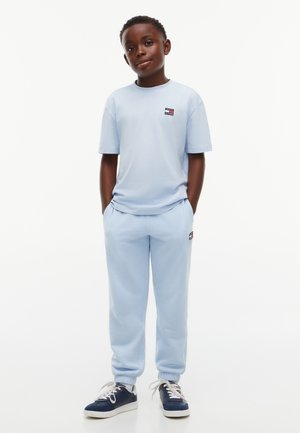 Garçon debout, mains dans les poches, portant un t-shirt bleu clair, un pantalon de survêtement et des baskets bleu foncé sur fond blanc.