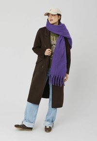 Écharpe oversize violette en matière douce, drapée élégamment autour du cou. Associée à un manteau marron foncé et un jean décontracté.
