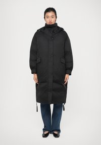 Long manteau en duvet noir avec un col haut, une fermeture à boutons à l'avant et deux poches latérales. Dispose de cordons de serrage à l'ourlet et aux poignets.
