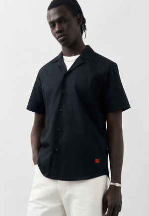 Young man with dark skin wearing a short-sleeve black button-up shirt over a white T-shirt and white pants, standing against a plain background.