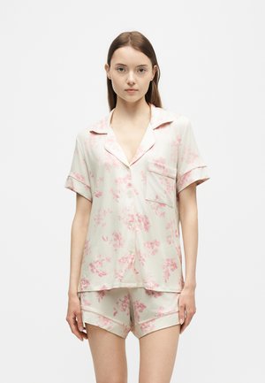 Jeune femme portant une chemise boutonnée à manches courtes crème et un short assorti avec des motifs floraux roses, debout devant un fond uni.
