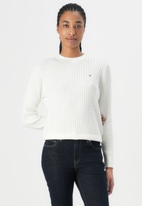 Hvit strikket genser med ribbestruktur, rund hals og lange ermer. Har en liten logo på brystet. Stylet med mørke jeans.