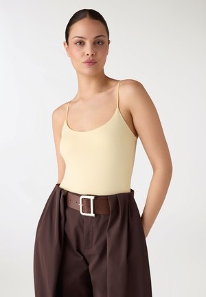 Donna con capelli scuri raccolti in uno chignon, che indossa una canotta giallo chiaro e pantaloni marrone scuro a vita alta con una cintura larga, in piedi davanti a uno sfondo bianco.