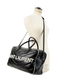 Sac à main en cuir noir Saint Laurent avec lettres blanches et passepoil, montré suspendu sur un torse de mannequin blanc.