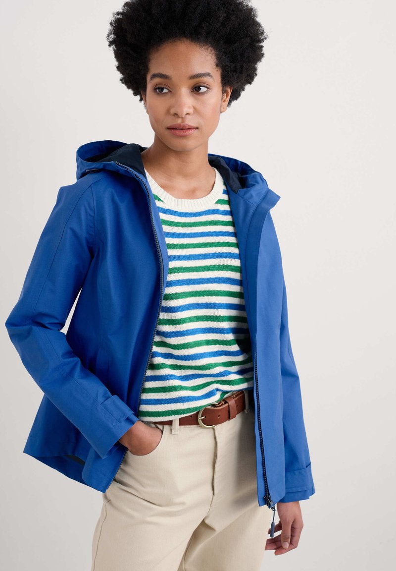 Mėlyna vandeniui atspari striukė su kapišonu, pasižyminti sklandžiu paviršiumi ir liekna forma. Užsidėta ant dryžuoto megztinio ir šviesiai rudos spalvos kelnių.