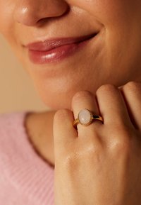 Goldring mit einem blassrosa ovalen Stein, umrandet von einem Perlenakzent. Der Hintergrund ist neutral und hebt die Details des Rings hervor.
