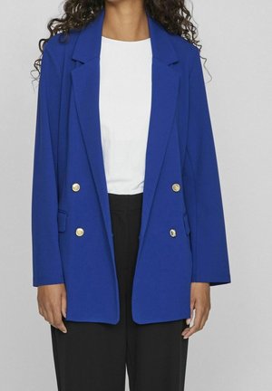 Blazer blu reale con design doppiopetto, bottoni dorati, revers a punta e tasche laterali, abbinato a una maglietta bianca e pantaloni neri.