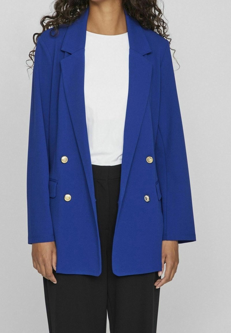 Královsky modrý blejzr s dvouřadým designem, zlatými knoflíky, klopami a bočními kapsami, v kombinaci s bílým topem a černými kalhotami.