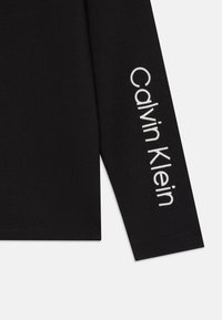 Crna majica dugih rukava od mekog materijala, s bijelim izvezenim tekstom "Calvin Klein" na lijevom rukavu. Jednostavan, prilagođen dizajn.