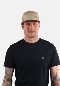 Casquette beige au design structuré et logo "OXBOW" brodé, portée avec un t-shirt noir arborant un petit motif blanc sur la poitrine.