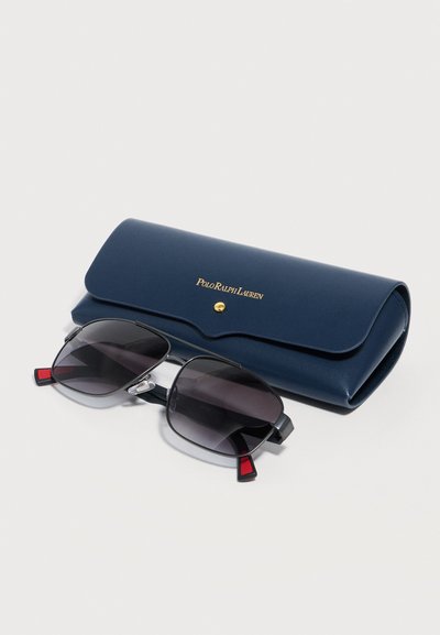 Μαύρα γυαλιά ηλίου με μαύρο περίγραμμα και σκουρόχρωνους φακούς που ακουμπούν μπροστά σε μια κλειστή σκούρα μπλε θήκη Polo Ralph Lauren με χρυσό κουμπί.