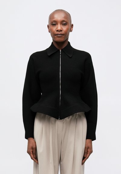 Black zip-up sweater made from textured fabric with a pointed collar and peplum hem, paired with light beige pleated trousers.