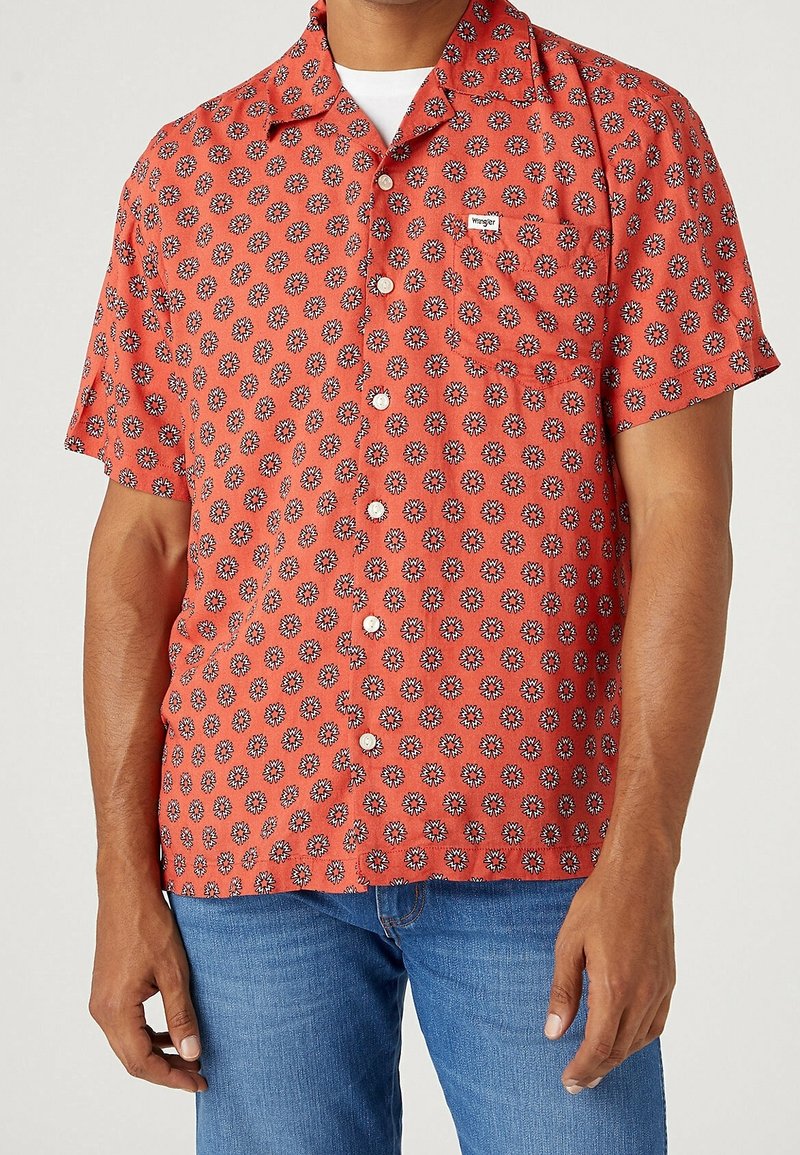 Homme portant une chemise corail à manches courtes avec un motif marguerite blanc et noir et un jean bleu, debout les bras détendus.