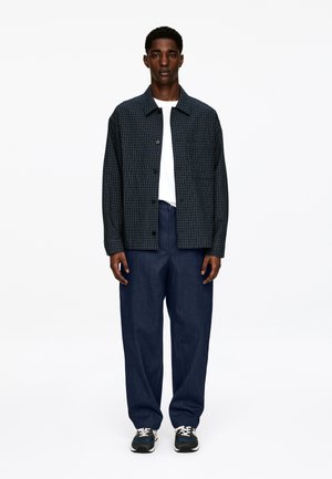 Checked navy jacket with dark green accents, white t-shirt underneath, paired with loose blue trousers, and dark sneakers.