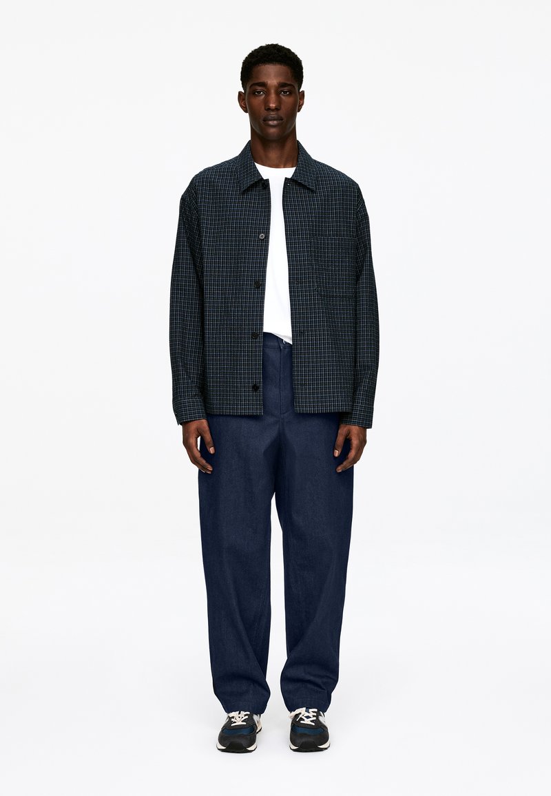 Ternet marineblå jakke med mørkegrønne detaljer, hvid T-shirt under, kombineret med løse blå bukser og mørke sneakers.