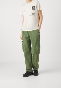 Camiseta beige de manga corta con un logo y texto en negro; pantalones cargo verdes con bolsillos con cremallera; calzado negro; tejido suave y corte relajado.