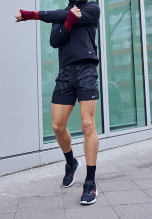 Persoon die zich uitstrekt, draagt een zwart sportjack, korte broek met patroon, zwarte sokken en zwarte sneakers, staande op een buitenpavement naast glazen ramen.