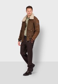 Giacca in suede marrone con colletto in pelliccia color crema, tasche frontali, dettagli con zip, indossata sopra una camicia verde oliva e pantaloni scuri.
