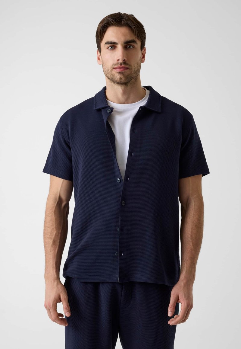 Homme aux cheveux courts et foncés portant une chemise bleu marine à manches courtes boutonnée par-dessus un T-shirt blanc, debout devant un fond clair uni.
