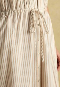 Ľahké šaty krémovej farby s vertikálnymi béžovými pruhmi. Majú opasok na viazanie, gombíkovú légou a jemné čipkované detaily okolo výstrihu.