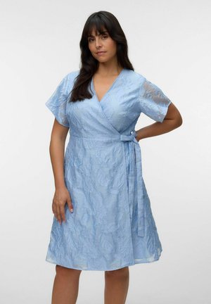 Kvinne som står med hånden på hoften iført en lyseblå teksturert omslagskjole med korte ermer og V-utringning på hvit bakgrunn.