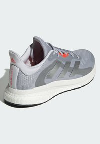 Šedá sportovní bota s prodyšným síťovaným svrškem, syntetickými překryvy, třemi texturovanými pruhy a bílou polstrovanou podrážkou. Obsahuje jasně oranžové akcenty.