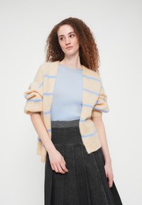 Πλεκτό ζακέτα σε μπεζ με μπλε ρίγες, με φαρδιά μανίκια και άνετη εφαρμογή. Συνδυασμένη με ανοιχτό μπλε τοπ και μαύρη πλισέ φούστα.