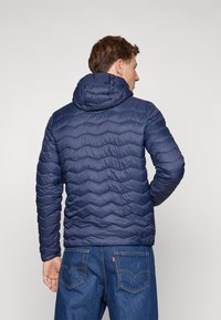 Veste matelassée bleu marine avec un design texturé et ondulé et une capuche. Associée à un jean en denim bleu, mettant en valeur un style décontracté et superposé.