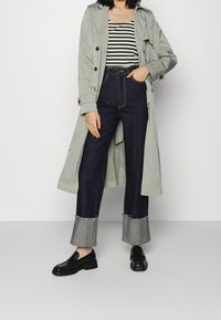 Vaaleanharmaa trenchcoat musta-valkoisen raidallisen paidan päällä, yhdistetty tummansinisiin leveisiin farkkuihin, joissa on purjehtimien reunat, ja mustiin mokkasiineihin.