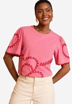 Roze T-shirt met korte mouwen, versierd met geborduurde rode cirkelpatronen en gegolfde randen, gecombineerd met lichtgekleurde broek.