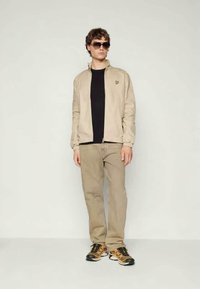 Gaiši brūns zipa jakas virs melna T-krekla, savienots ar gaiši brūnām biksēm un daudzkrāsainiem sporta apaviem. Iekļautas saulesbrilles un minimāla zīmolu identificēšana.