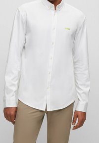 Camicia bianca button-up con maniche lunghe, logo verde chiaro sul petto e bottoni verde chiaro. Tessuto liscio e design del colletto classico.