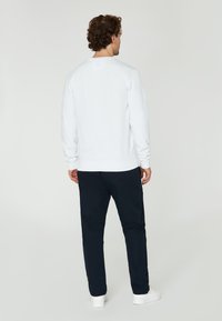 Sudadera blanca con cuello redondo, puños y dobladillo acanalados, combinada con pantalones oscuros y zapatillas blancas, vista desde la espalda.