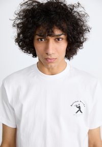 White cotton t-shirt with short sleeves, featuring a small black graphic print on the left chest area depicting a figure with a trumpet.