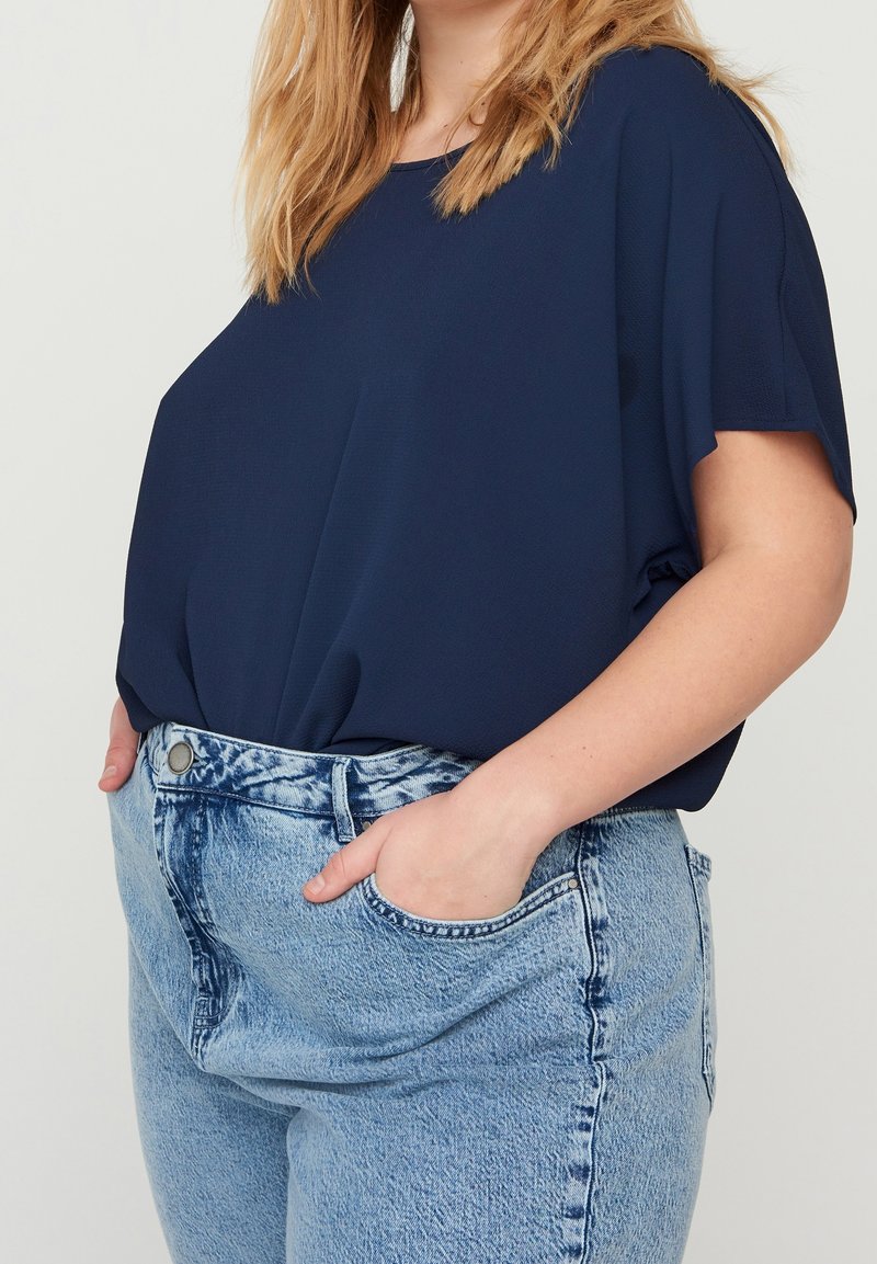 Femme portant un haut ample bleu marine à manches courtes rentré dans un jean taille haute bleu clair délavé, les mains dans les poches avant.